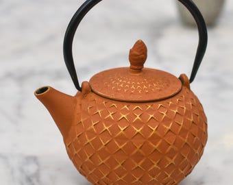 May include: A terracotta-colored teapot with a black handle and a textured, diamond-patterned surface. The lid is topped with a small, cone-shaped knob. The teapot has a spout for pouring and is designed for brewing tea.