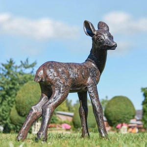May include: A bronze deer statue stands on grass against a blue sky. The deer has a detailed, textured surface and is posed with its head turned. This decorative sculpture is a garden ornament.