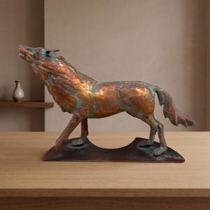 May include: A copper sculpture of a wolf in a howling pose, featuring a weathered patina with visible teal and green oxidation accents. The wolf stands on a dark, curved metal base, showcasing a textured surface with metallic highlights.
