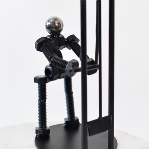 May include: A black metal sculpture of a person made of nuts and bolts holding a cricket wicket. The sculpture is on a black base.