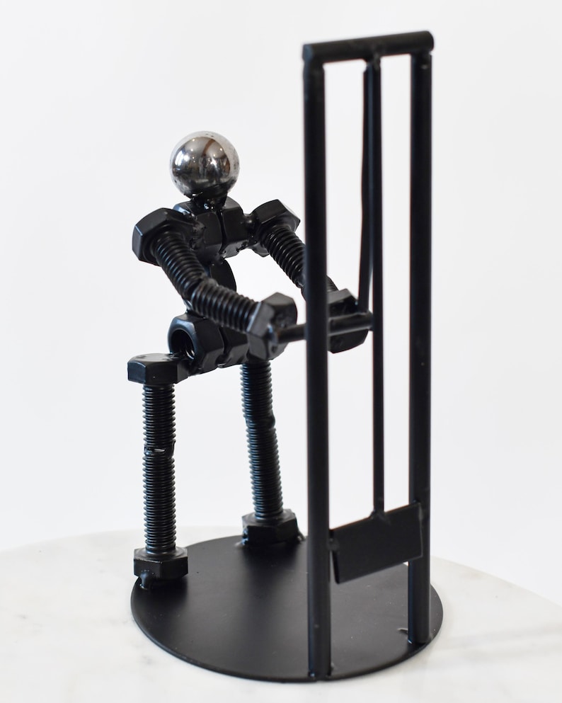 May include: A black metal sculpture of a person made of nuts and bolts holding a cricket wicket. The sculpture is on a black base.