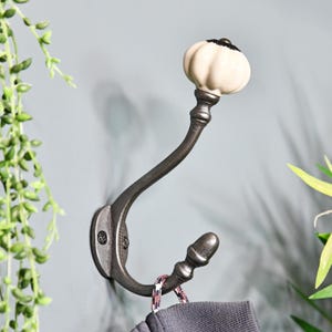 May include: A cast iron double hook with a white ceramic knob. The hook is mounted on a wall and has a grey finish.