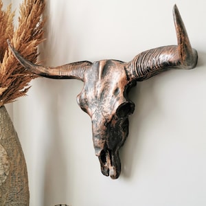 May include: A wall-mounted bull skull sculpture featuring a metallic copper and black finish. The piece displays detailed bone texture, prominent curved horns, and a hollow nasal cavity, creating a striking contrast against the light wall.