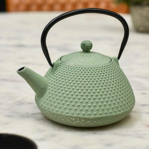 May include: A sage green cast iron teapot with a black handle and spout. The teapot has a textured surface with small, raised dots. The lid has a small knob. The teapot is on a marble surface.
