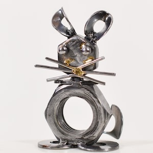 May include: A metal sculpture of a mouse, made from nuts, bolts, and washers. The mouse has upright ears, whiskers, and a tail. The body is formed from a large nut with a circular opening. The sculpture is silver with gold accents.