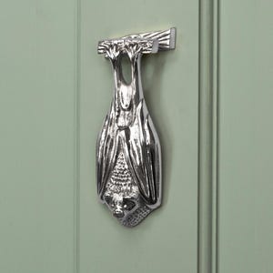 May include: A polished silver bat door knocker, mounted on a light green door. The bat is depicted hanging upside down, with its wings neatly folded. The metal knocker features intricate detailing, adding a decorative touch to the door.
