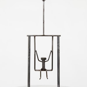 May include: A metal sculpture depicting a person on a swing. The figure is constructed from a bolt and metal rods, with a sphere atop a vertical rod. The sculpture is mounted on a round base. The overall height is approximately 20 cm.