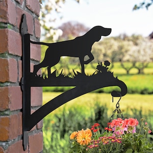 May include: Black metal silhouette of a dog with a hanging planter. The planter is filled with colourful flowers. The silhouette is mounted on a brick wall.