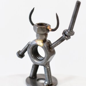 May include: A handcrafted metal sculpture of a figure made from nuts, bolts, and screws. The figure has a nut for a body, a bolt for an arm, and a screw for a horn. The sculpture is on a white background.
