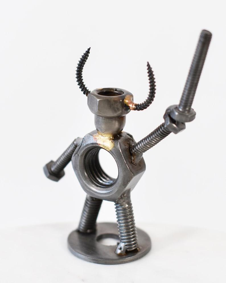 May include: A handcrafted metal sculpture of a figure made from nuts, bolts, and screws. The figure has a nut for a body, a bolt for an arm, and a screw for a horn. The sculpture is on a white background.