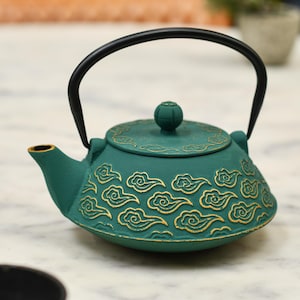May include: A teal cast iron teapot featuring a raised gold-coloured cloud pattern across the body and lid. The teapot has a curved black metal handle attached to the top and a small spout on the left side, with a round knob on the centre of the lid.