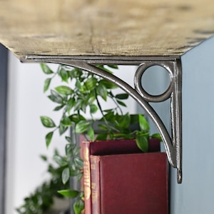 May include: A rustic wooden shelf with a black metal bracket. The bracket has a decorative ring design and is attached to a wall.