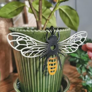 May include: A metal bee decoration with white wings and a black body, featuring a yellow and orange striped abdomen. The bee is attached to a green ceramic plant pot.