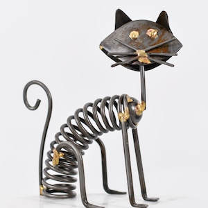 May include: A metal cat sculpture with a spring body, upright ears, and a long tail. The cat's head and legs are made of metal, with gold-coloured accents. The sculpture is a unique decorative item.