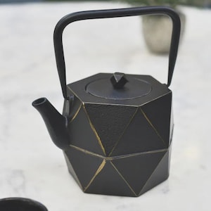 May include: Black geometric teapot with a handle and spout. The teapot has a lid with a small knob and gold accents. The teapot is on a white surface. A black teacup is in the foreground.