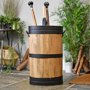 May include: A wooden umbrella stand with black metal detailing. The cylindrical stand features a light brown wood grain finish and black bands. Several umbrellas and walking sticks are inside the stand. Green plants are in the background.
