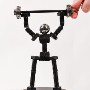 May include: A black metal sculpture of a person lifting a barbell. The sculpture is made of nuts and bolts and is standing on a black base.