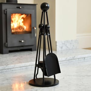 May include: A black metal fireplace tool set featuring a circular base, a central stand with spherical finials, a shovel, a poker, and a brush. The tools are arranged vertically within the frame, showcasing a matte finish and a sturdy construction.