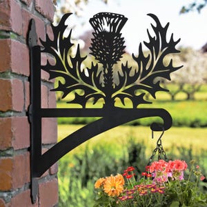 May include: Black metal garden bracket with a thistle design. The bracket is attached to a brick wall and has a hanging chain with a flower pot.