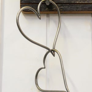 May include: A silver metal heart-shaped wall hook. The hook features two intertwined hearts. It is mounted on a dark wooden board and attached to a white wall. The hook is designed for hanging items.