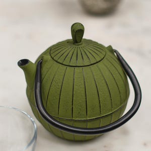 May include: A green cast iron teapot with a black handle and spout. The teapot has a ribbed design and a small knob on the lid. A clear glass cup is in the foreground, and a silver cup is in the background.