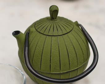May include: A green cast iron teapot with a black handle and spout. The teapot has a ribbed design and a small knob on the lid. A clear glass cup is in the foreground, and a silver cup is in the background.