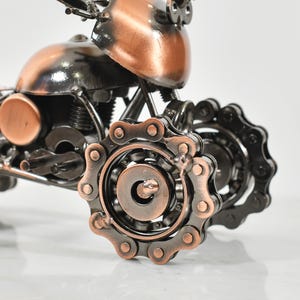 Puede incluir: Una peque&ntilde;a escultura de motocicleta de metal hecha con piezas recicladas. La motocicleta es de color negro y cobre y tiene una transmisi&oacute;n por cadena.
