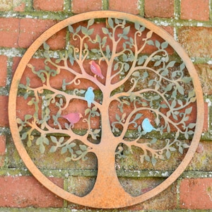 May include: A circular metal tree of life wall decoration with a rusty brown finish. The tree design includes cut-out leaves and branches, with four colourful birds perched on the branches. It is mounted on a red brick wall.