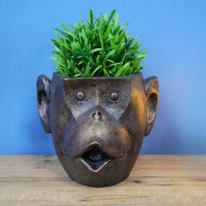 May include: A bronze-effect monkey-shaped plant pot with a wide open mouth and large ears. The pot is filled with vibrant green artificial grass. The pot is on a wooden surface with a blue background.