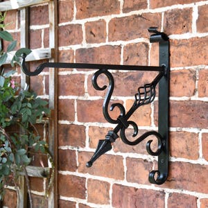 May include: Black metal wall-mounted plant hanger with a decorative scroll design. The hanger has a single arm extending out from the wall with a hook at the end.