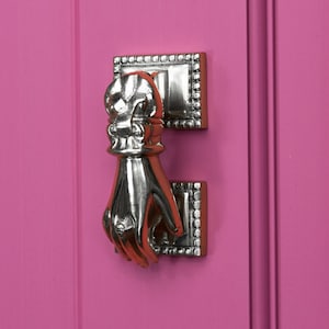 May include: A silver door knocker shaped like a hand with a decorative top. The knocker is mounted on a pink door.