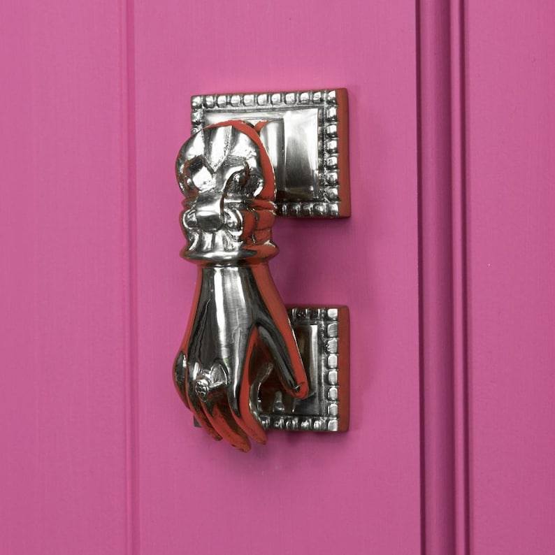 May include: A silver door knocker shaped like a hand with a decorative top. The knocker is mounted on a pink door.