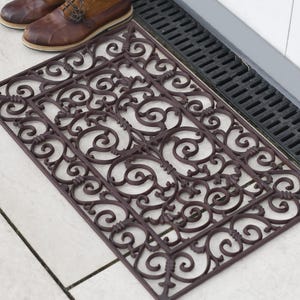 May include: A brown, decorative doormat with a scrolled design. The doormat is made of rubber and has a textured surface.