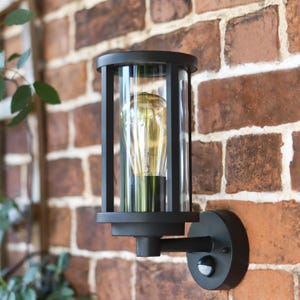 May include: Black outdoor wall light with a cylindrical glass shade and a decorative cage. The light features a vintage-style bulb and is mounted on a red brick wall. The fixture includes a motion sensor.