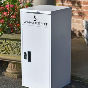 May include: A white, rectangular post box with a hinged lid. The text "5 ASHMOLE STREET" is printed in black on the front. A black combination lock and handle are on the door. The post box is outdoors, near a brick wall and flowers.