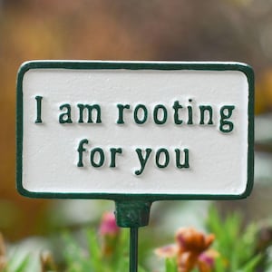 May include: A white rectangular garden marker with the words "I am rooting for you" in dark green. The marker has a dark green border and is placed in a garden with green plants and flowers.