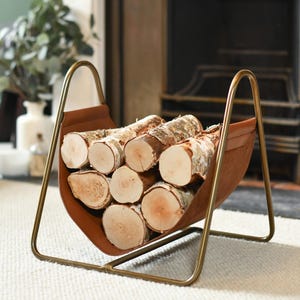 May include: A brown leather and brass log holder filled with birch logs. The holder has a curved brass frame and a leather sling to hold the logs. The logs are cut into rounds and have a light-coloured wood grain.