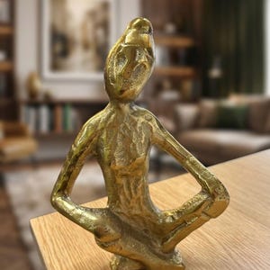 May include: A gold-coloured sculpture of a seated figure with arms resting on knees. The figure has a textured surface and a stylized, abstract design. The sculpture is placed on a light-coloured wooden surface.