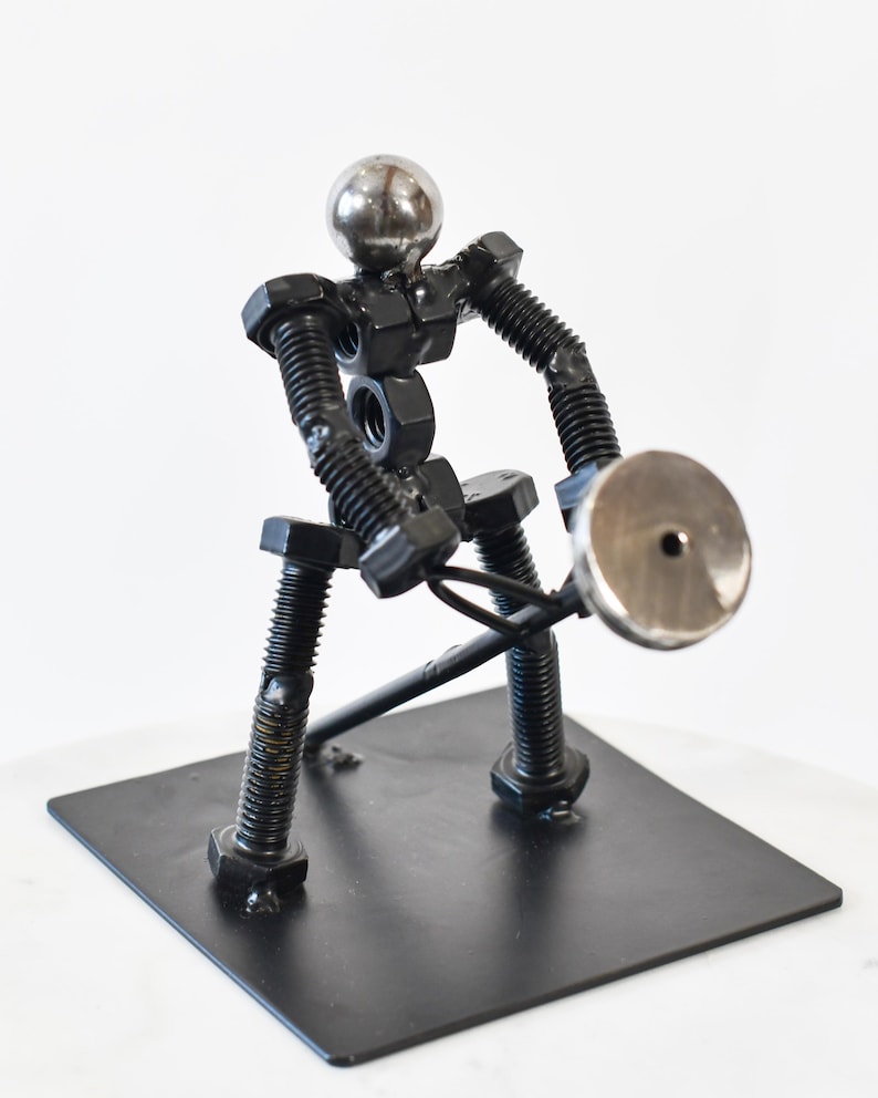 May include: A metal sculpture of a person lifting a weight. The sculpture is made of nuts, bolts, and other metal pieces. The person is standing on a black metal base.