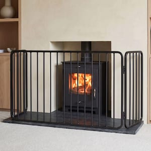 May include: A black metal fireplace guard with a multi-panel design and vertical bars, positioned in front of a black wood-burning stove with a visible fire inside. The guard features rounded corners and a sturdy frame structure.