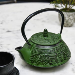 May include: Green cast iron teapot with a black handle and lid. The teapot features an intricate embossed design. A black teacup is visible in the foreground. The teapot is designed for brewing tea.