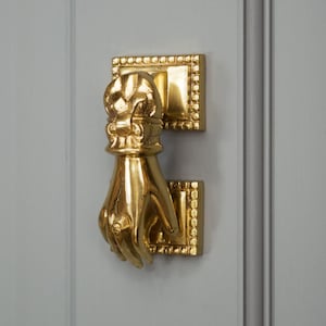 May include: A gold-toned door knocker shaped like a hand with a decorative design on the back of the hand. The knocker is mounted on a rectangular base with a decorative border.