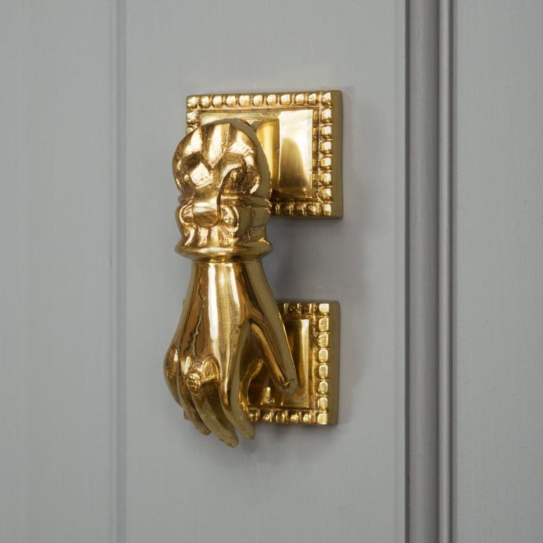 May include: A gold-toned door knocker shaped like a hand with a decorative design on the back of the hand. The knocker is mounted on a rectangular base with a decorative border.