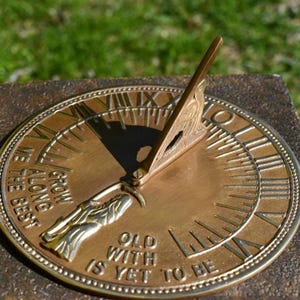 May include: A circular brass sundial featuring a central gnomon and Roman numerals engraved around the perimeter. The surface includes the embossed text "GROW OLD ALONG WITH ME THE BEST IS YET TO BE" and a small figure holding a scythe.