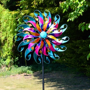 May include: A vibrant metal garden ornament in the shape of a flower. The design incorporates a central blue disc and curved petals in pink, orange, and teal. It is mounted on a black pole, set against a backdrop of green foliage.