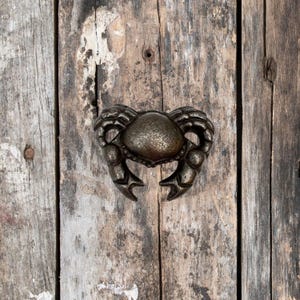 May include: A cast iron crab bottle opener with a rustic brown finish. The opener is attached to a wooden surface.