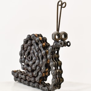 May include: A metal snail sculpture crafted from recycled bicycle chain, nuts, and bolts. The snail has a coiled body, a head with nut eyes, and antennae. The metal has a dark, aged finish, giving it a rustic appearance. The sculpture is a unique piece of art.