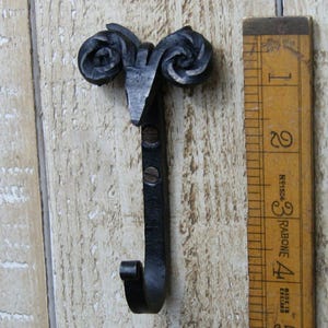 May include: A handcrafted, black metal hook with a ram's head design. The hook is approximately 10 cm long and features a curved hook end. The ram's head has detailed spiral horns. The hook is mounted on a wooden surface.
