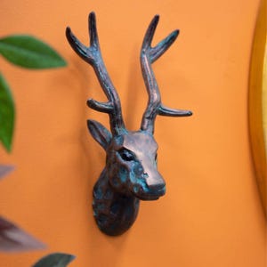 May include: A bronze deer head wall sculpture with a blue-green patina finish. The sculpture is mounted on an orange wall. The antlers and facial features are clearly visible. This artwork adds a rustic touch to home decor.