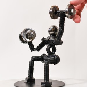 May include: A metal sculpture of a person lifting weights. The sculpture is made of nuts, bolts, and washers. The person is sitting on a round metal base.